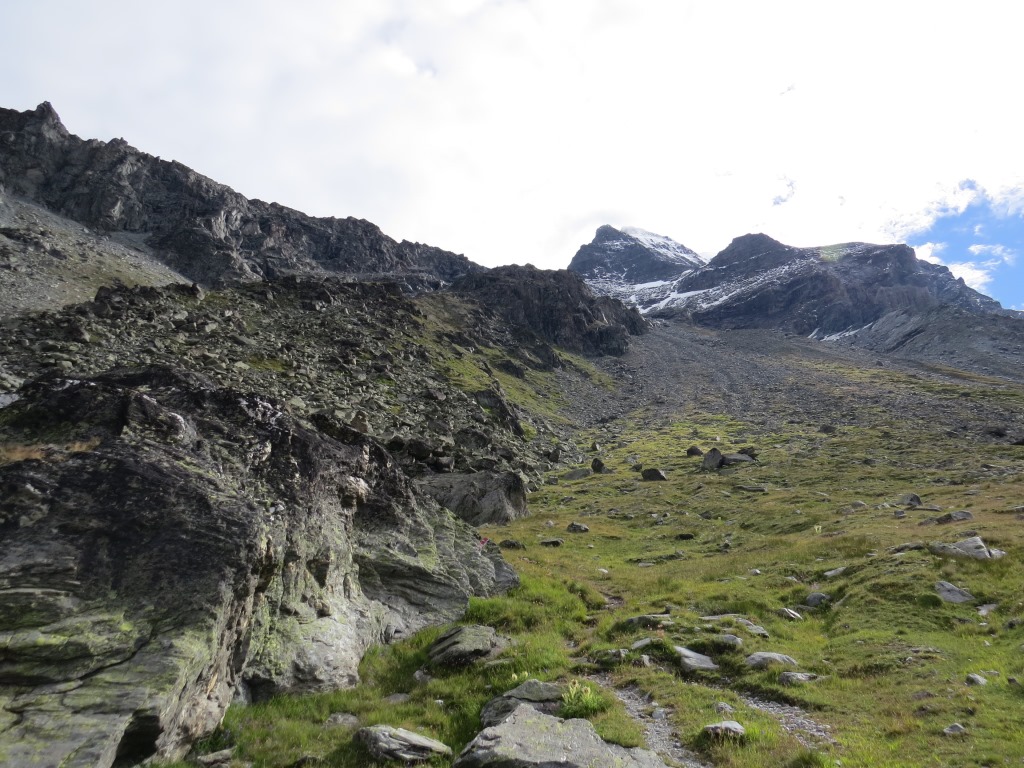 über die Wiese mit einigen Felsblöcken führt der Weg auf die Rippe zu, auf der hoch droben die Hütte zu sehen ist